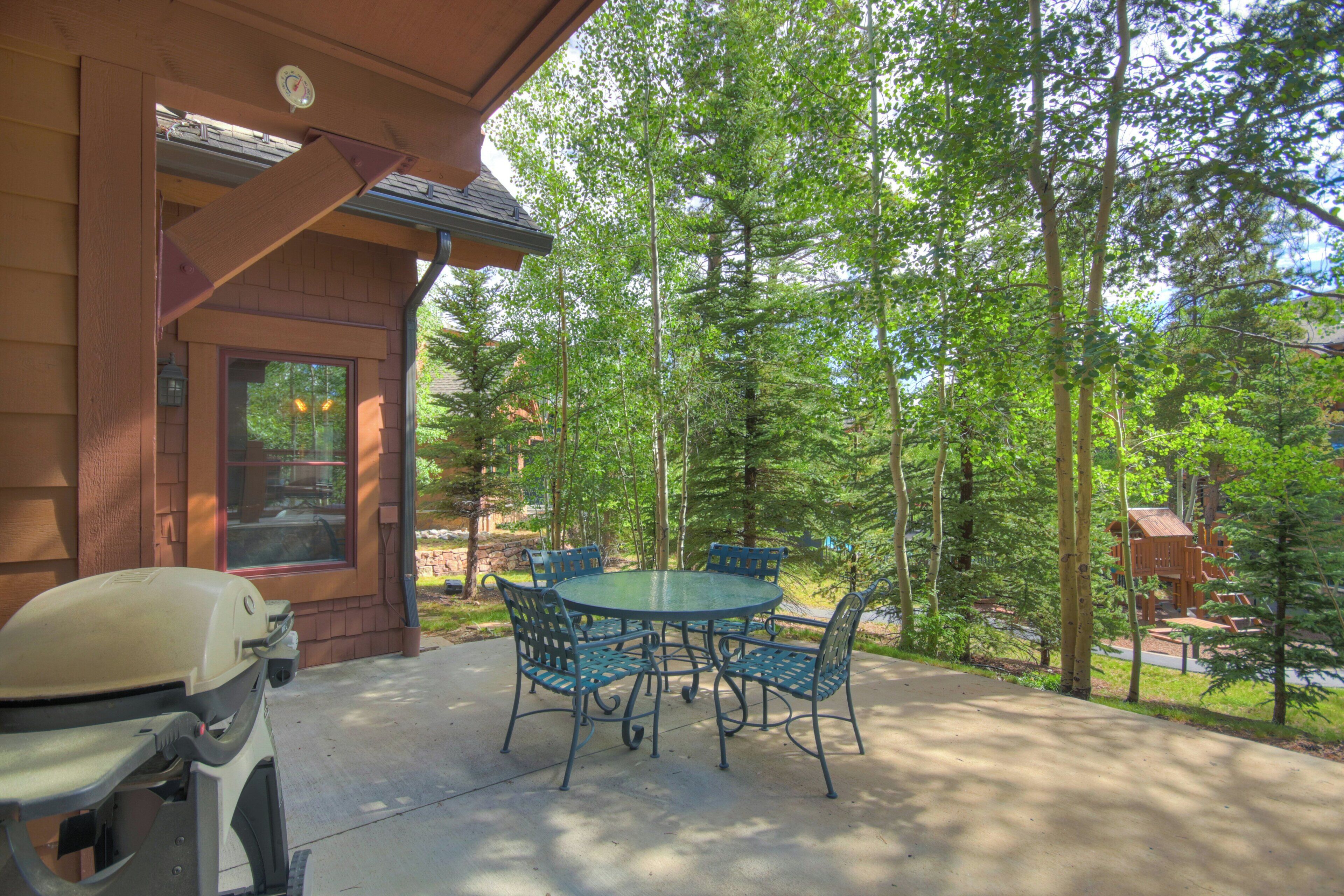 A patio with a round table and chairs, a barbecue grill, and a view of a forested area.
