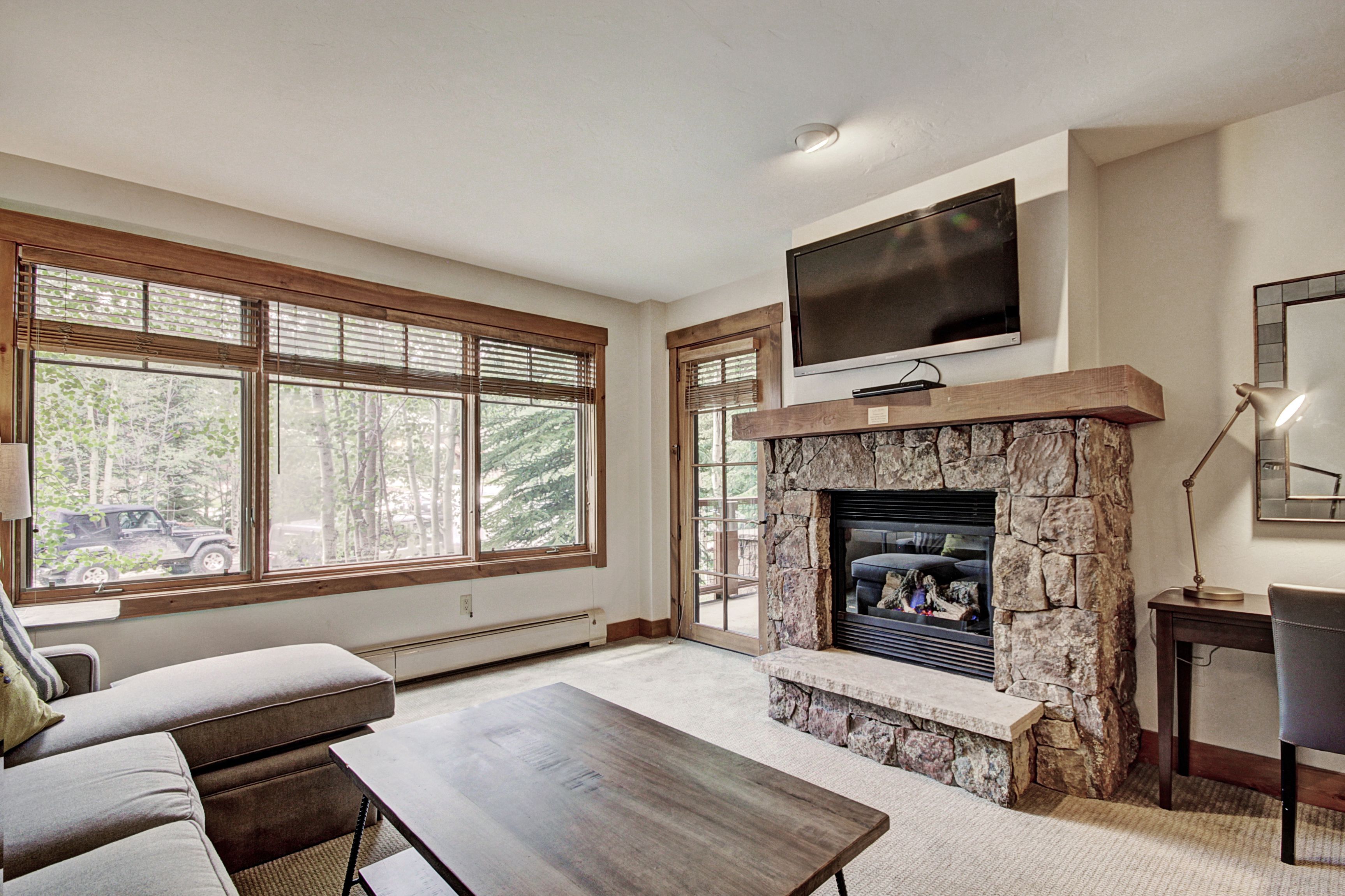 A spacious living room with a stone fireplace, a flat-screen TV mounted above it, and large windows offering a view of the outdoors.