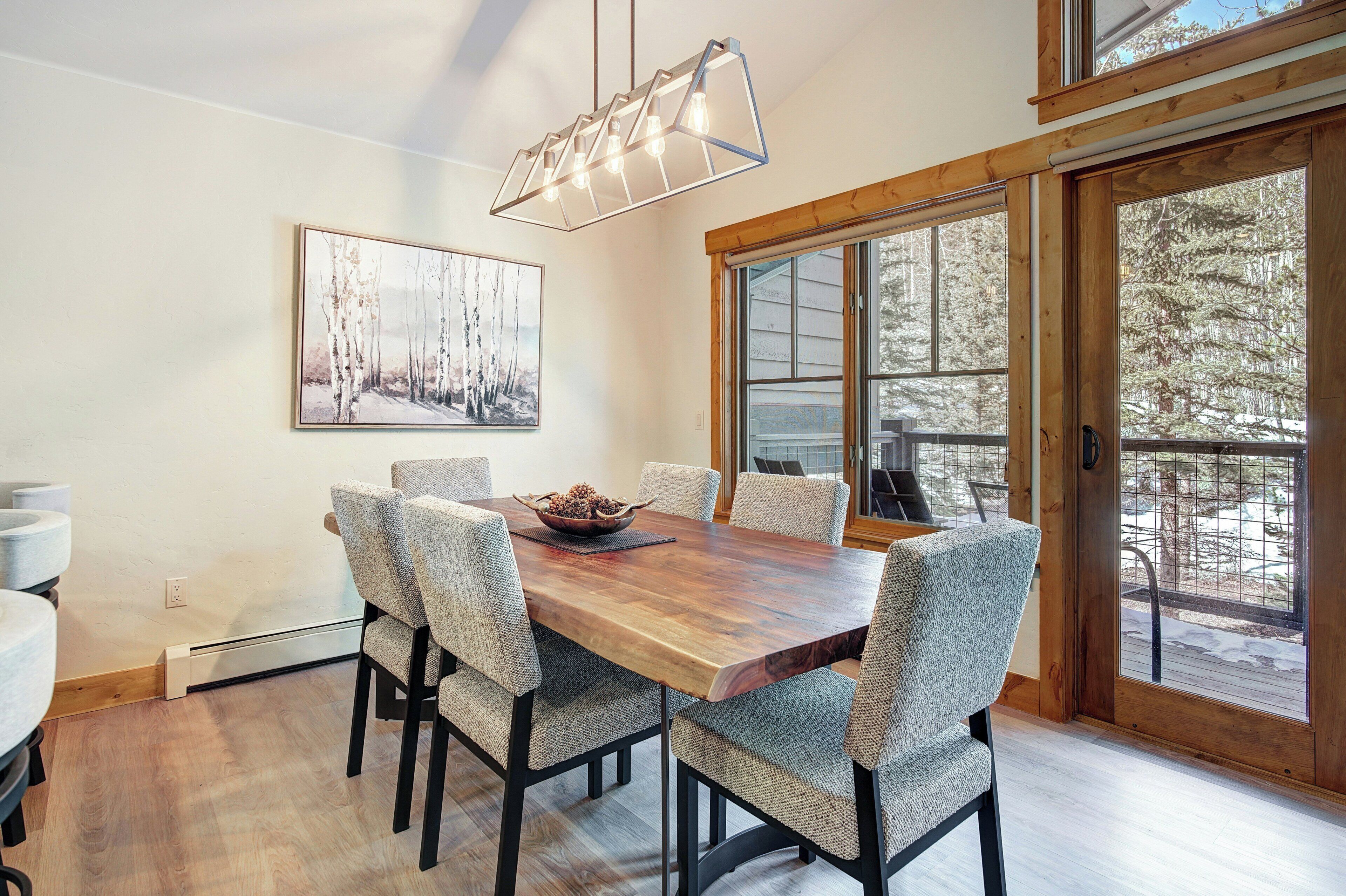 A dining area with a wooden table, six chairs, a hanging light fixture, and a large window with a view of trees.
