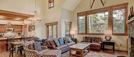 A spacious living area with a high ceiling, a fireplace, and large windows offering a view of the outdoors.
