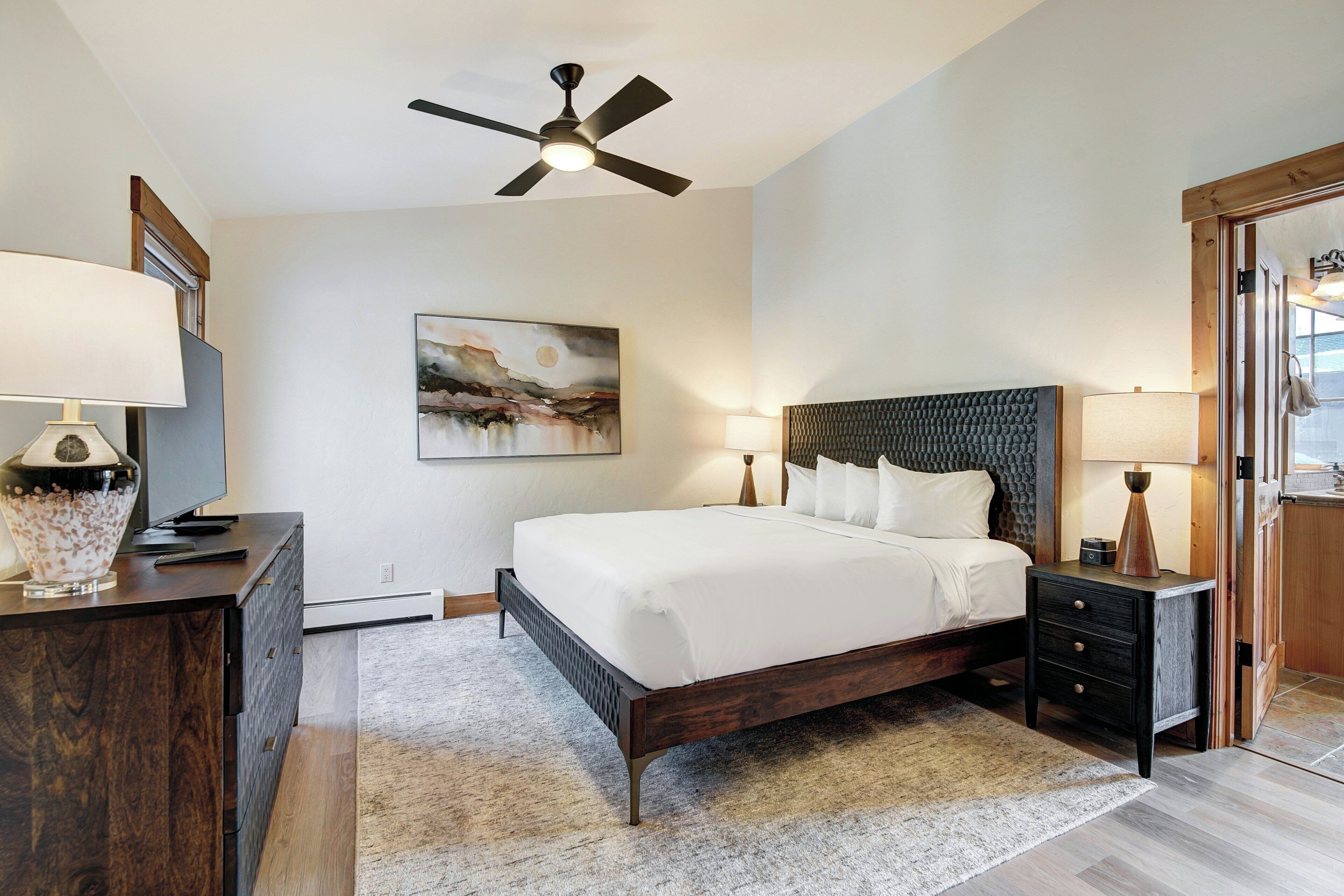 A bedroom with a bed, bedside tables, a ceiling fan, a painting, and a dresser.