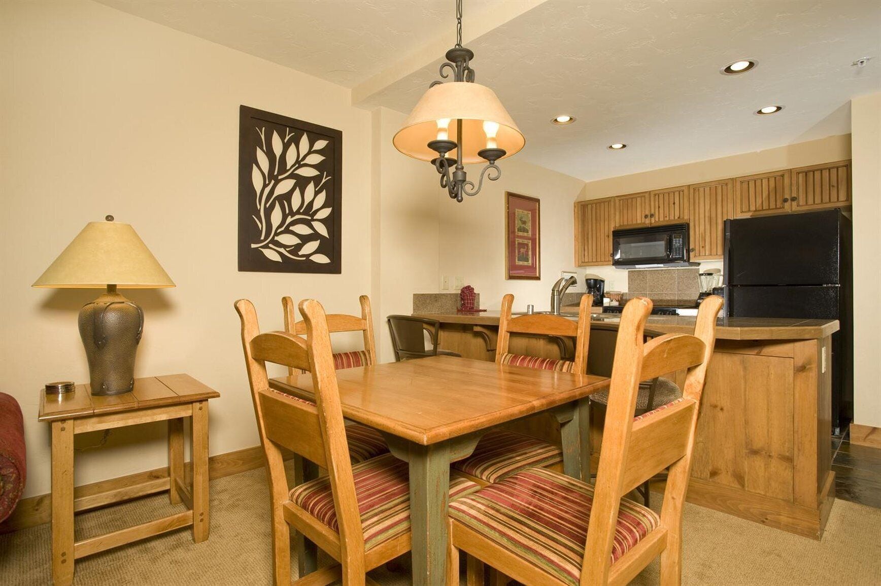 A dining area with a wooden table and chairs, a hanging lamp, and a kitchen in the background.