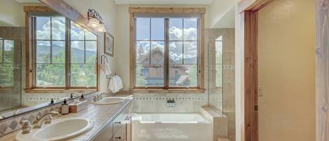 A spacious bathroom with two sinks, a large bathtub, and a window offering a view of a mountain and a cabin.
