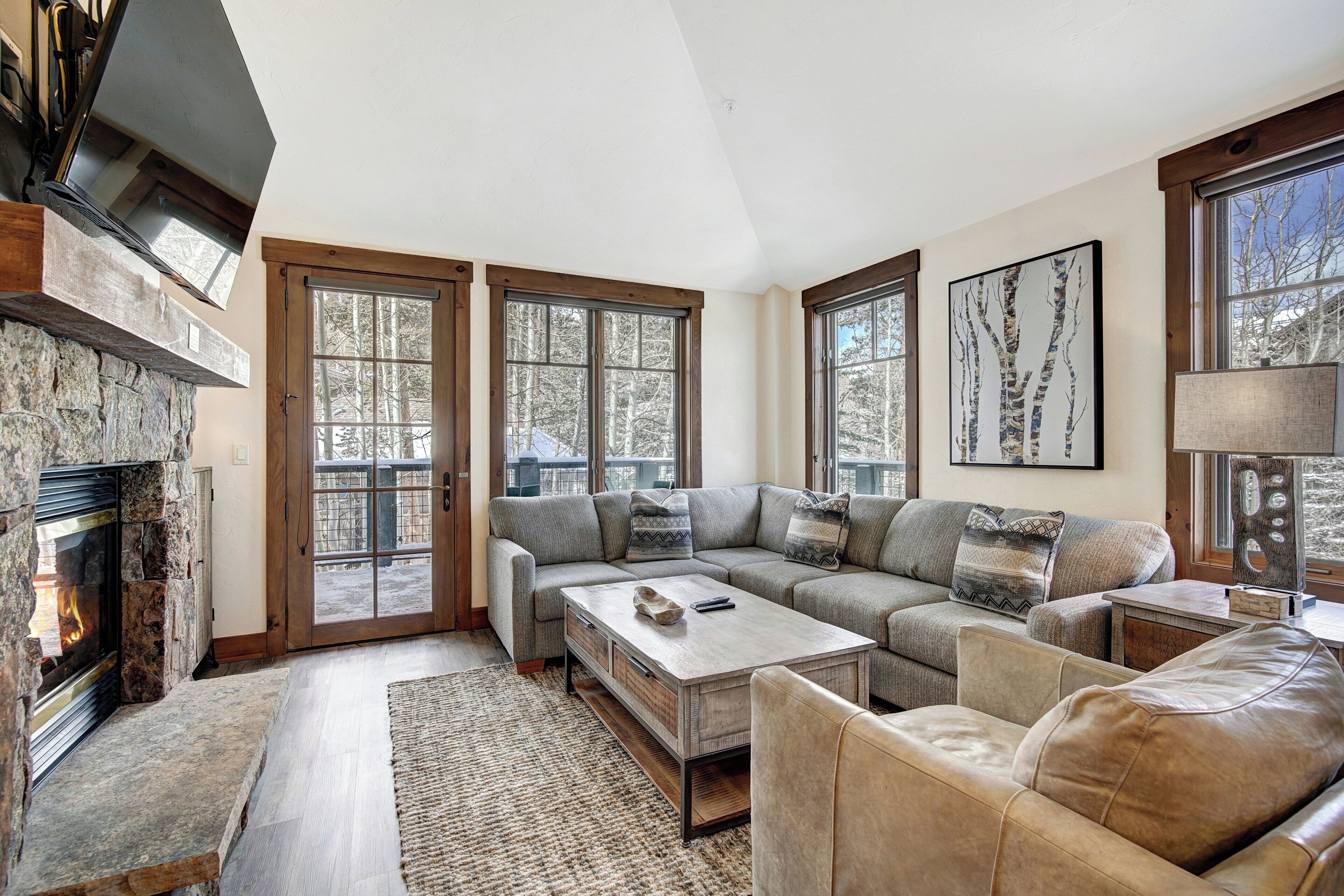 A living room with a stone fireplace, a sectional sofa, a coffee table, and large windows with a view of trees.