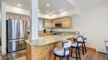 A modern kitchen with a stainless steel refrigerator, wooden cabinets, a granite countertop, and bar stools.