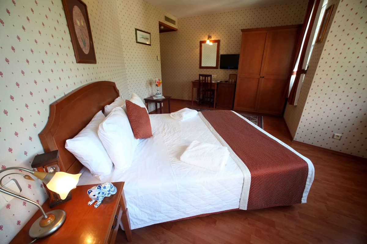 A hotel room with a large bed, a desk, a chair, a TV, and a wardrobe.