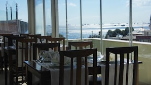 A restaurant with tables set for dining, large windows offering a view of the sea and distant buildings.