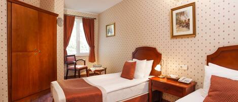 A hotel room with two beds, a wooden wardrobe, a desk with a lamp, and a window with curtains.