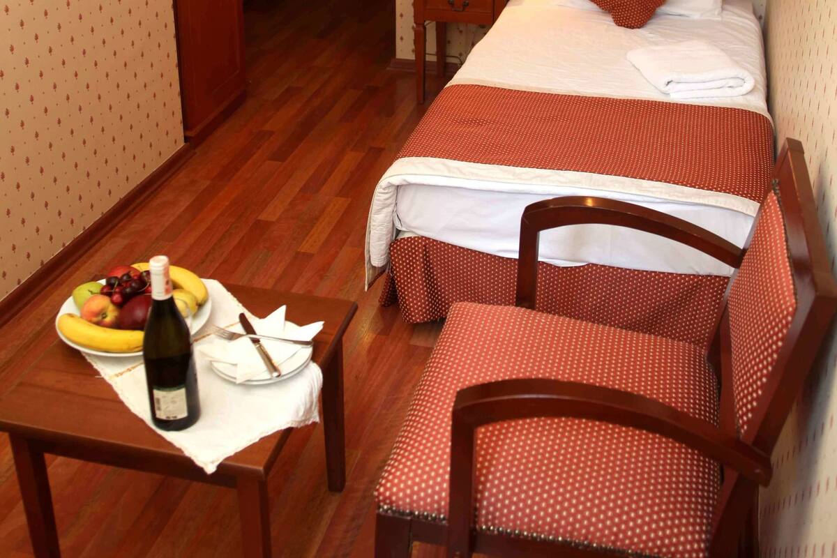 A hotel room with a bed, a bedside table with a lamp, a chair, and a small table with a bottle and fruit.