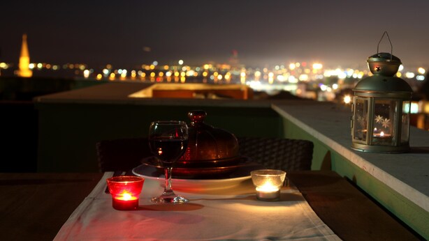 Une table avec une bougie, un verre de vin et une lanterne, le tout installé sur un toit offrant une vue sur la ville illuminée la nuit.