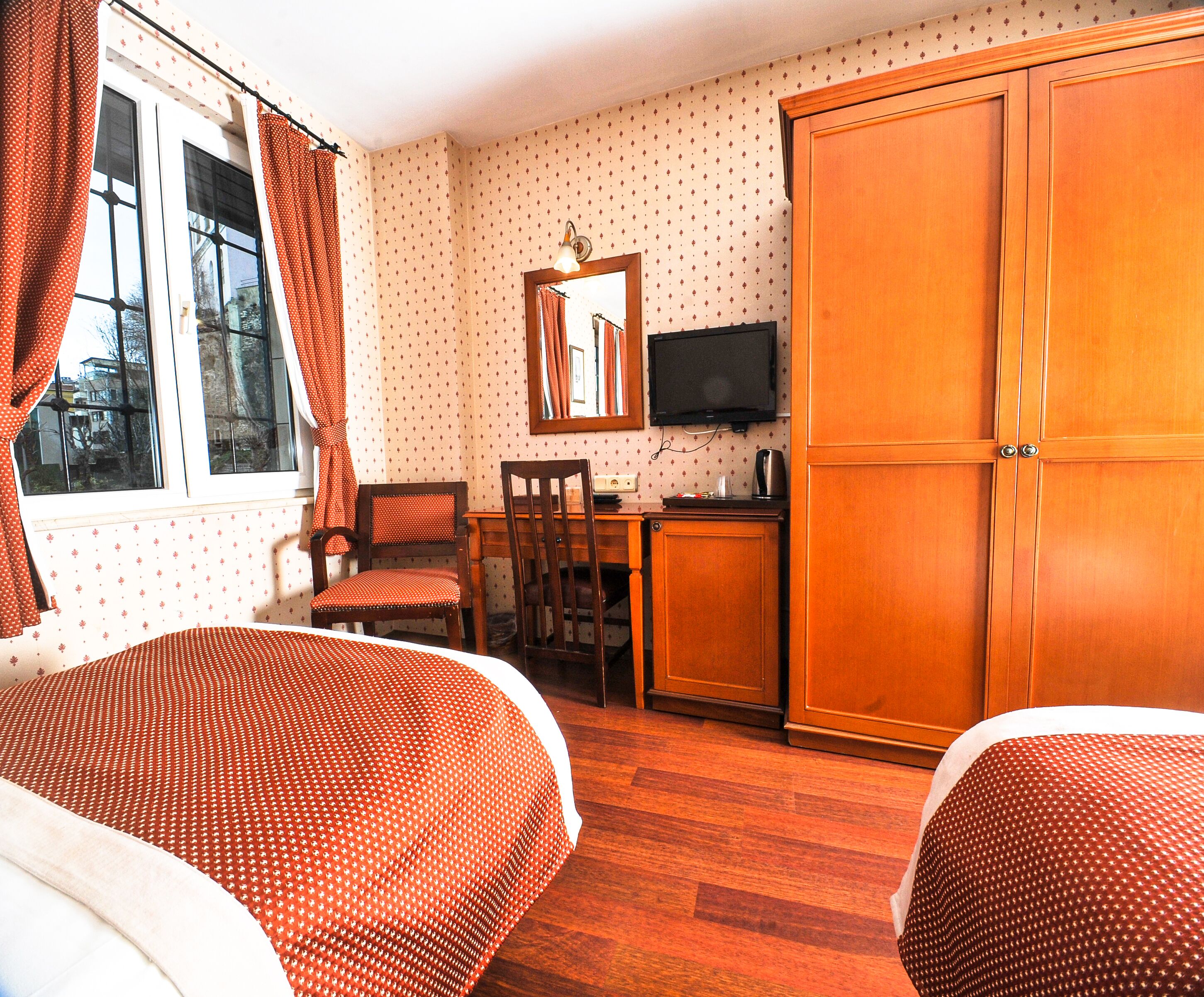 A hotel room with two beds, a desk with a chair, a wardrobe, a television, and a window with curtains.