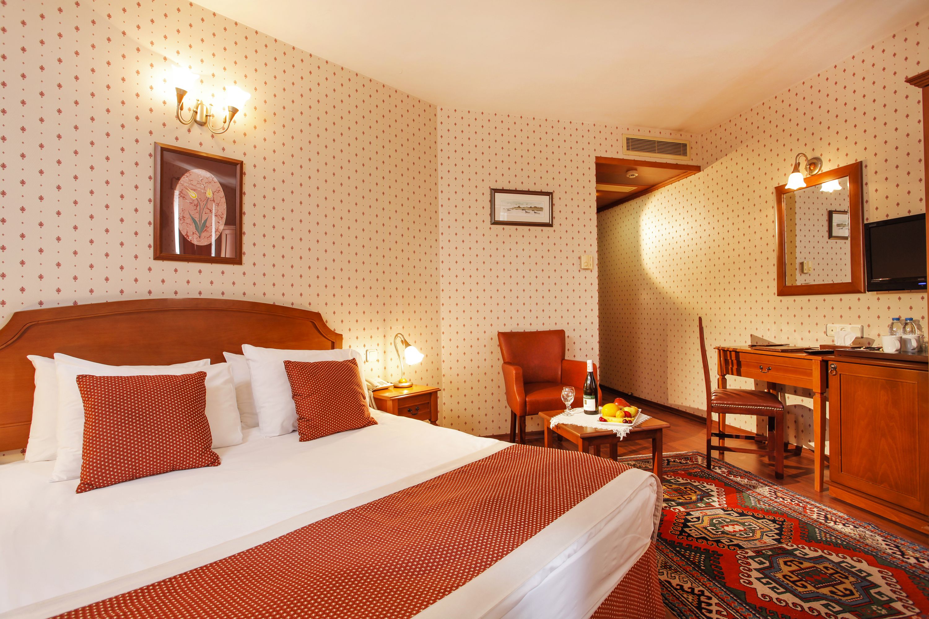 A hotel room with a large bed, a wooden headboard, a desk with a chair, a television, and a patterned rug.