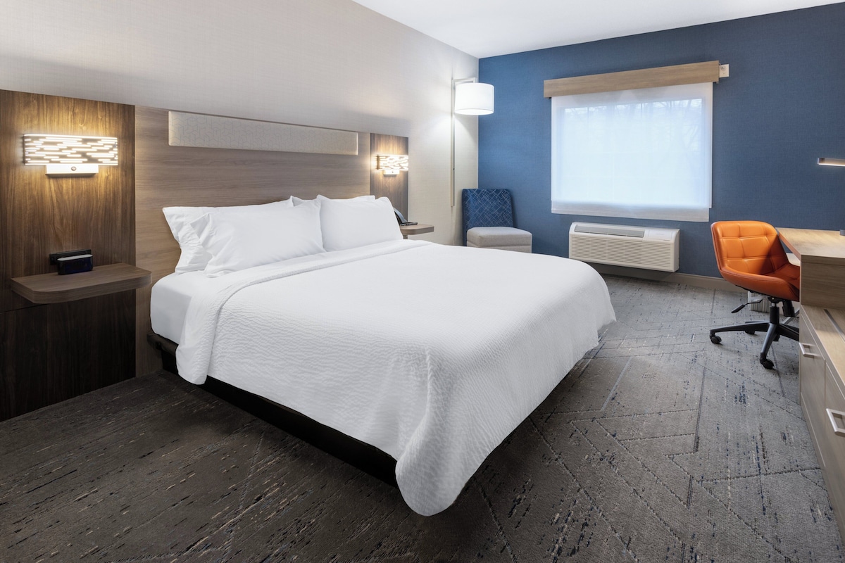 A hotel room with a large bed, bedside tables, a desk with an orange chair, a radiator, and a window with curtains.