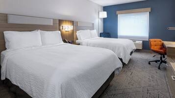 A hotel room with two beds, a desk, an orange chair, a nightstand with a lamp, and a window with blinds.