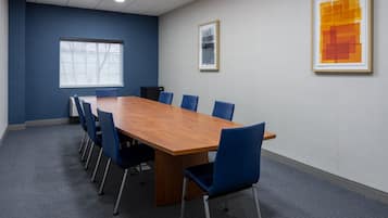 Ein Konferenzraum mit einem langen Holztisch, blauen Stühlen, einem großen Fenster und zwei gerahmten Kunstwerken an den Wänden.