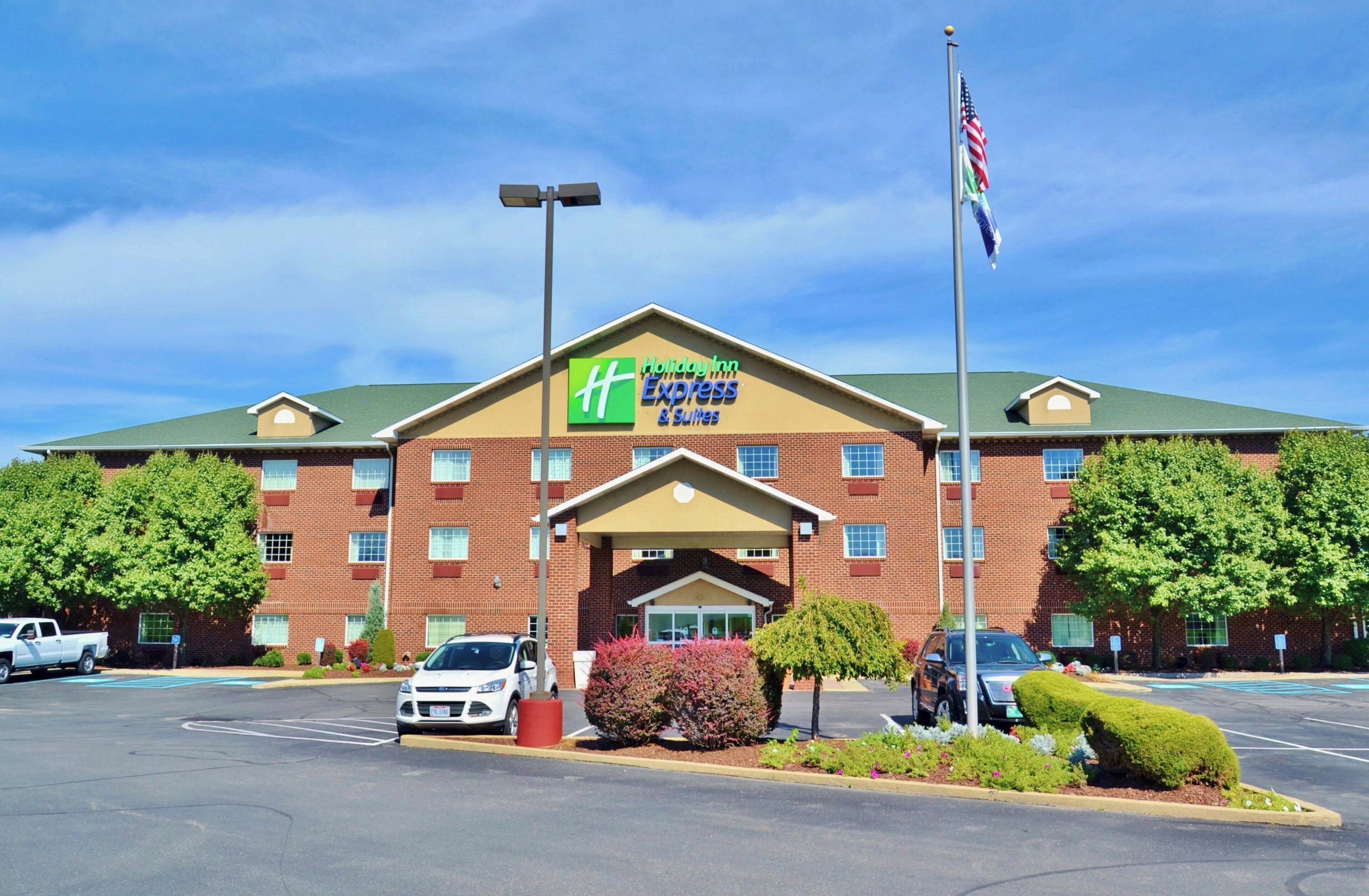 Un hotel Holiday Inn Express con fachada de ladrillo, techo verde y un estacionamiento con varios coches.