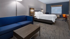 A hotel room with a bed, a blue sofa, a wooden table, a lamp, and a window. - Holiday Inn Express & Suites Center Township by IHG (Monaca)