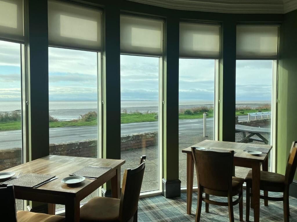 Comedor con grandes ventanales que ofrecen vista al mar y a una carretera. Las mesas están preparadas con platos y cubiertos.