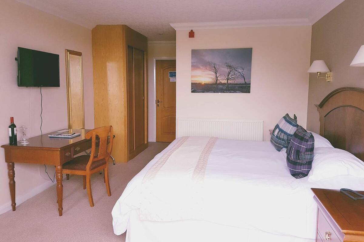 A hotel room with a bed, a desk with a chair, a television, a painting on the wall, and a lamp.