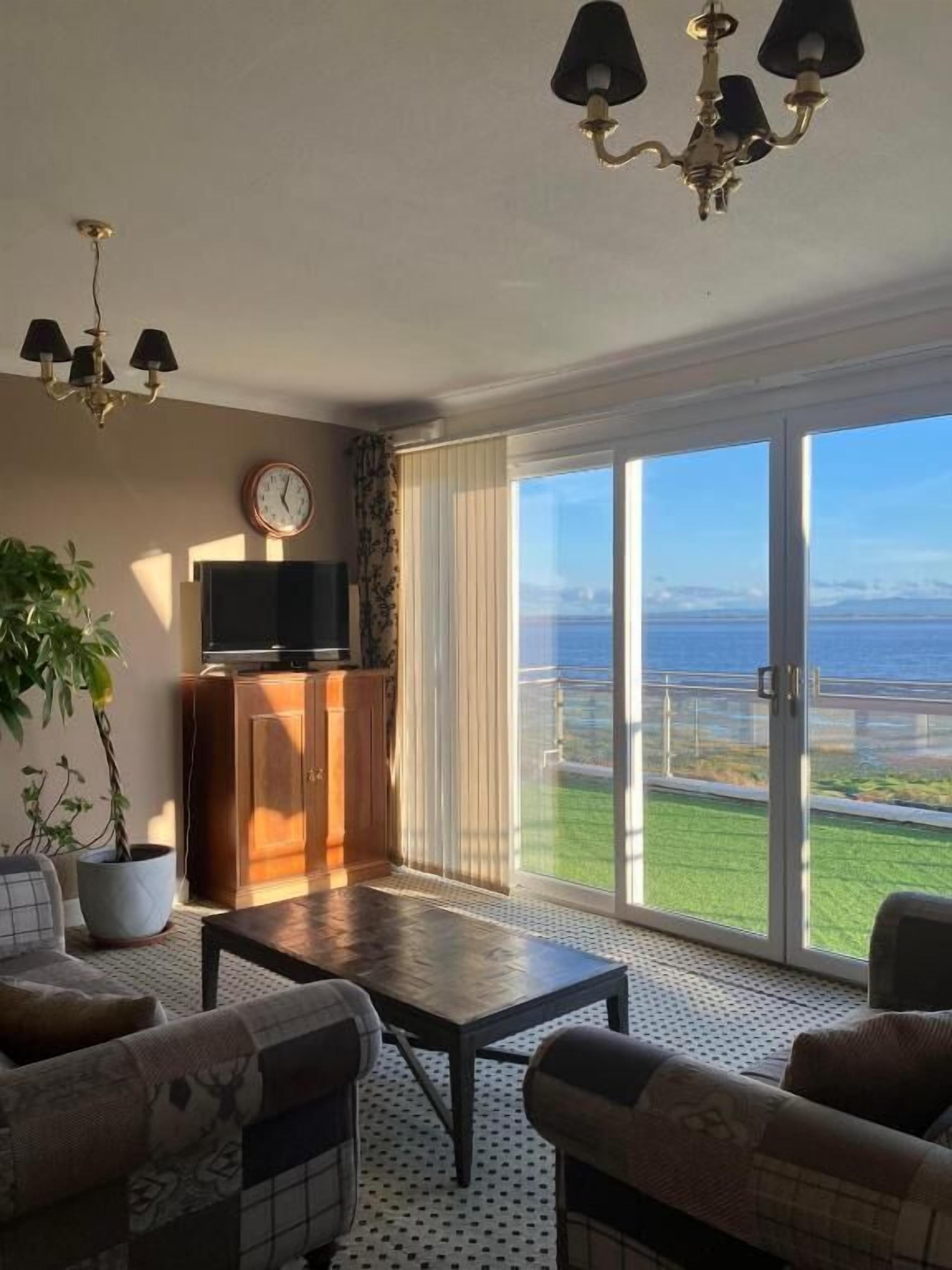 Un salón con sofá, mesa de centro y televisión sobre un mueble. Se ve una ventana grande con vistas al mar.
