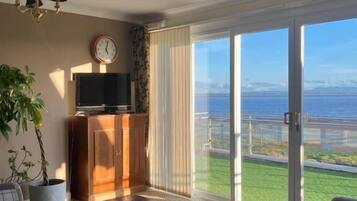 Un salón con sofá, mesa de centro y televisión sobre un mueble. Se ve una ventana grande con vistas al mar.