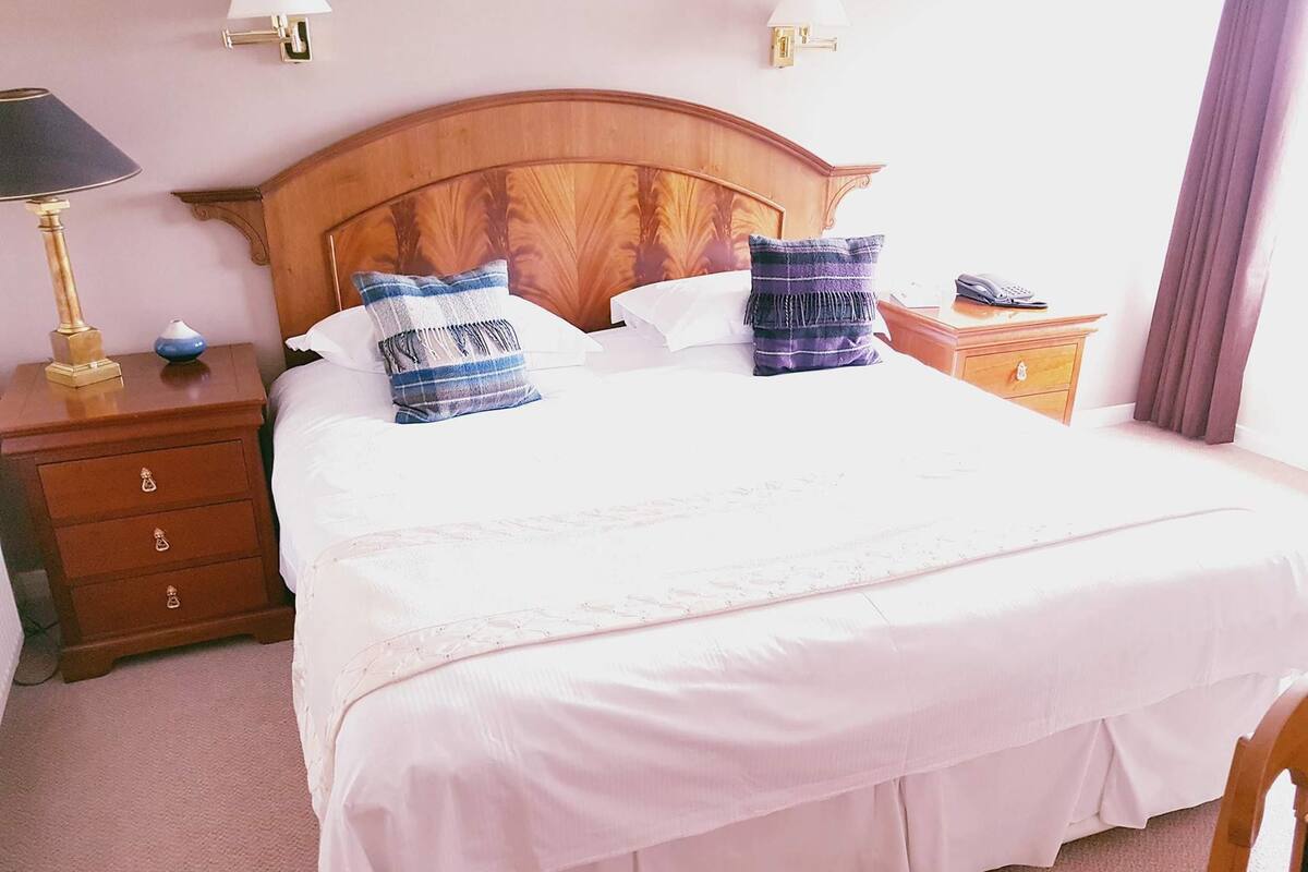 A neatly made bed with white linens and two decorative pillows, a wooden nightstand with a lamp and a vase, and a bedside table with a phone.