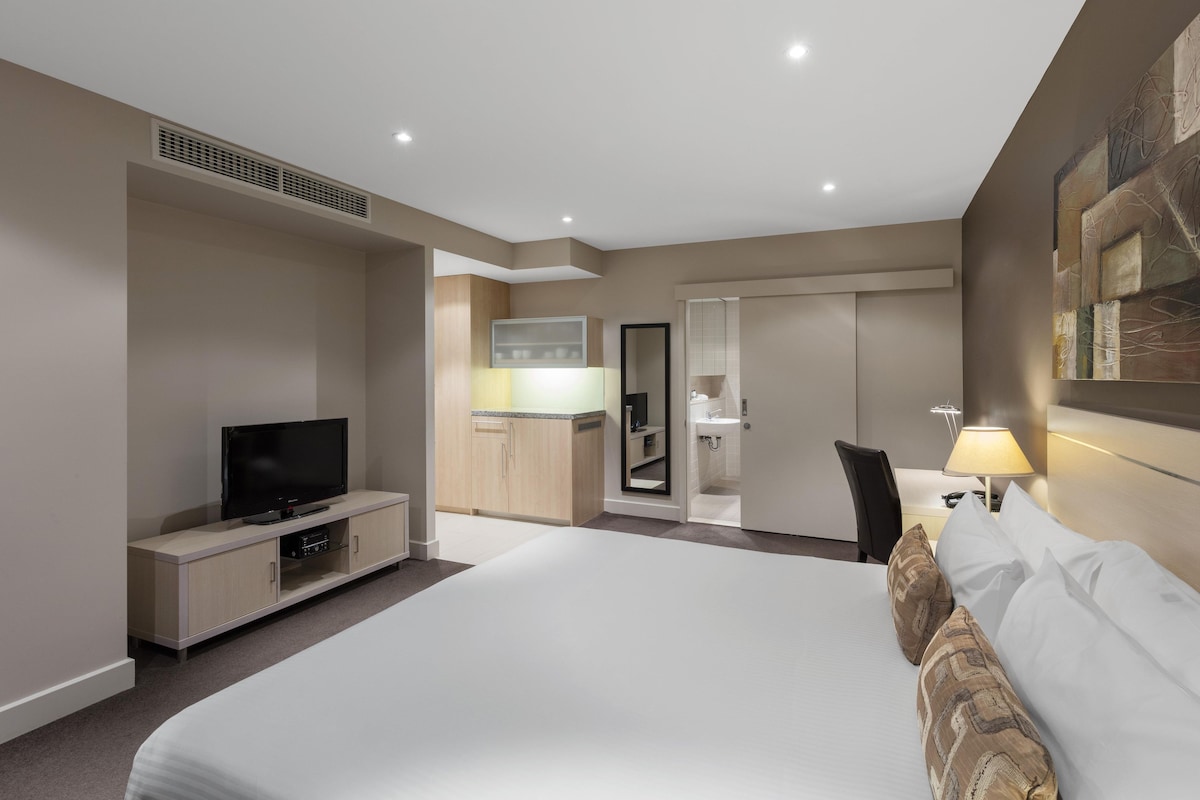 A modern hotel room with a large bed, a flat-screen TV, a desk, and a bathroom visible through an open door.