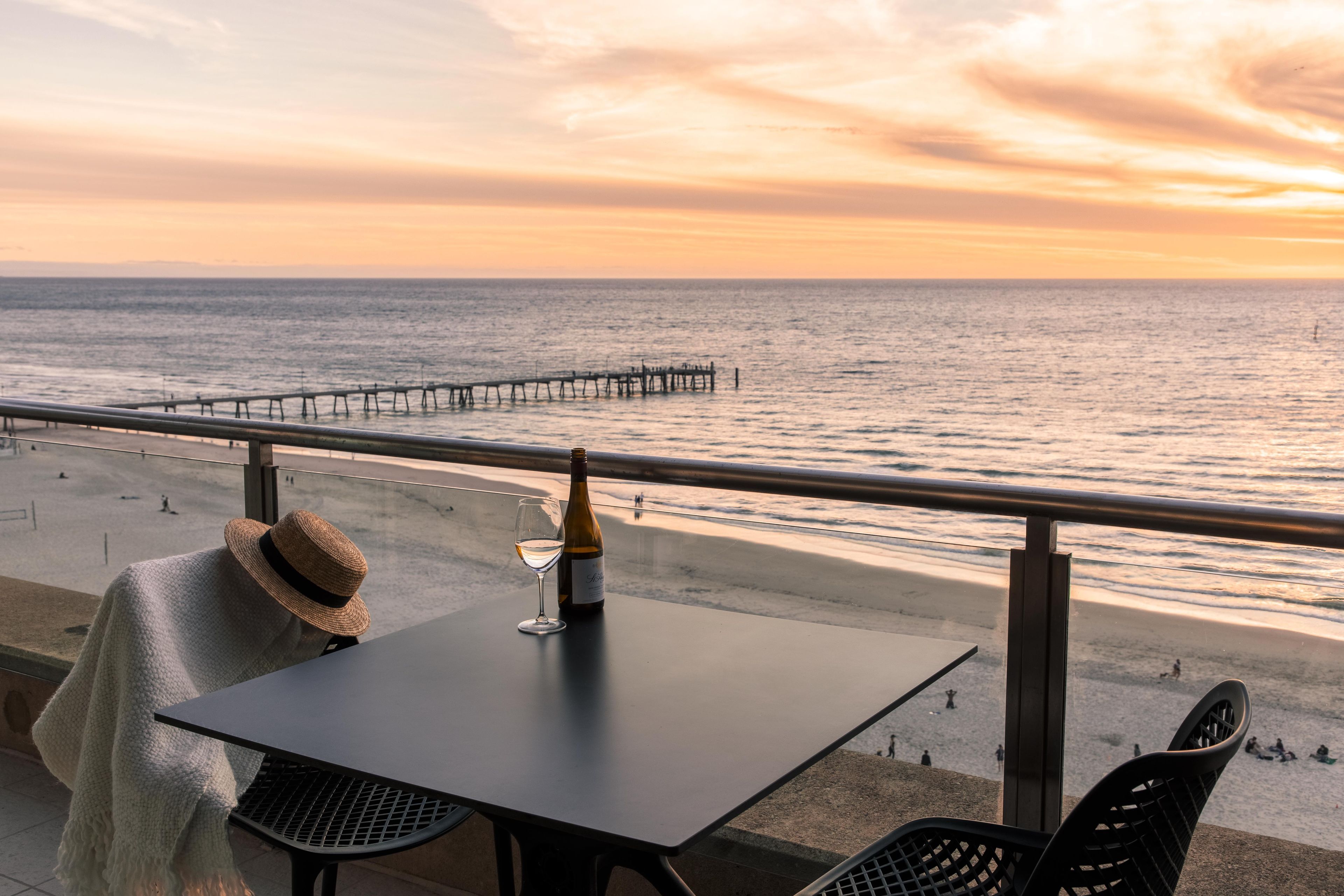 A table with a bottle and glass, a straw hat, and a view of the beach and ocean at sunset.