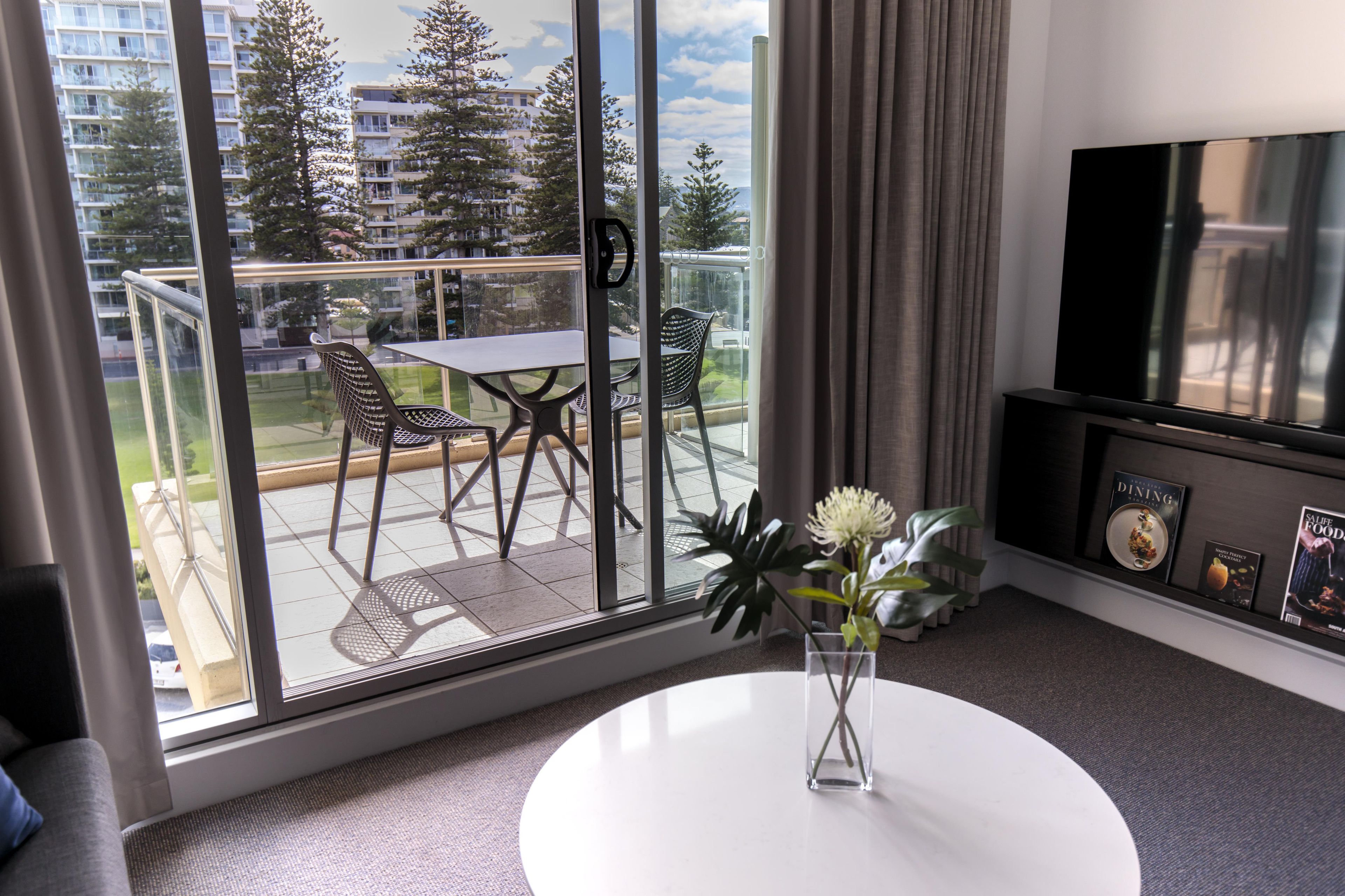 A modern living room with a large window, a round white table, and a flat-screen TV.
