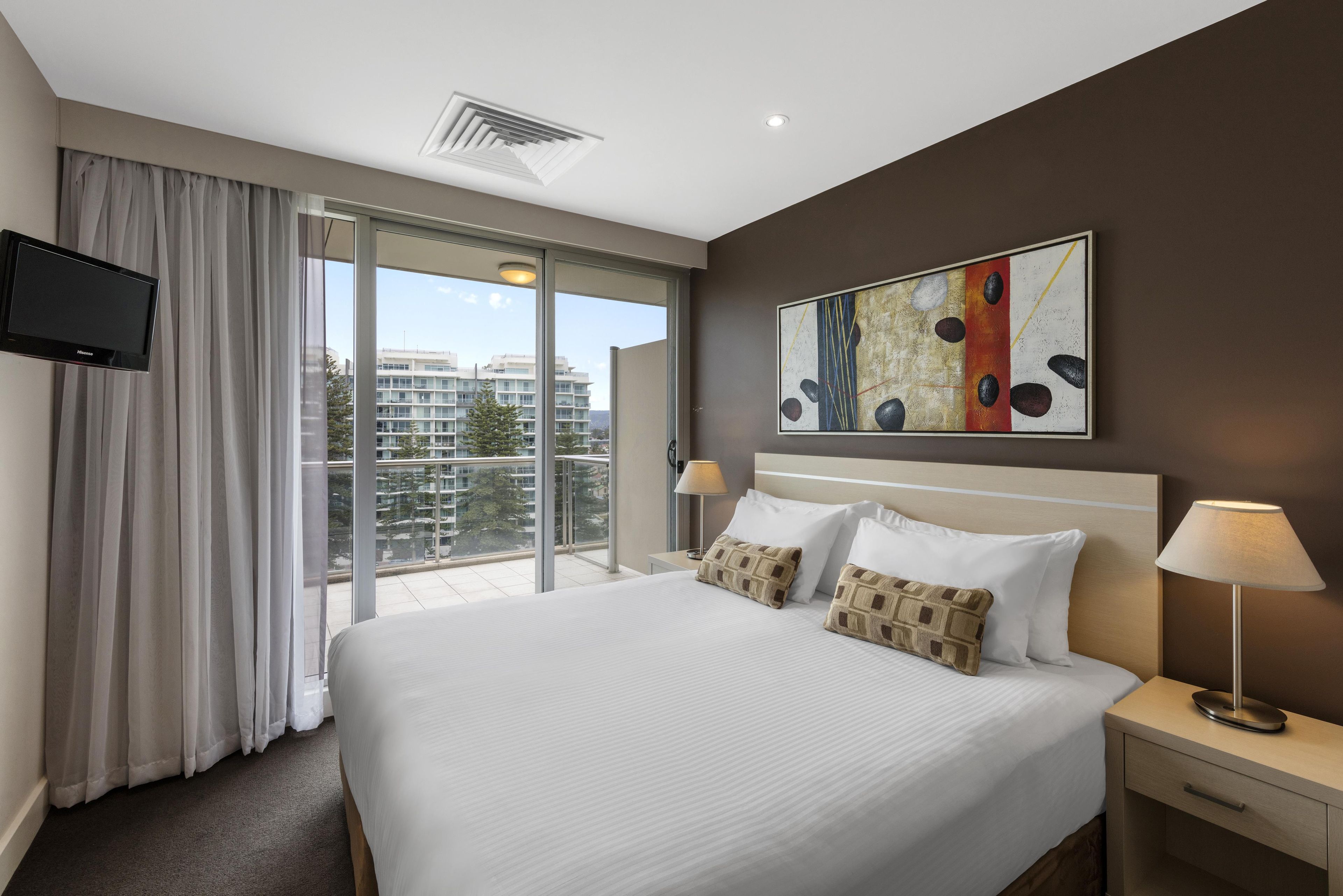 A hotel room with a bed, a flat-screen TV, a painting, a nightstand, a lamp, and a view of buildings through the window.