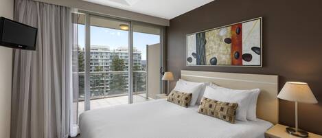 Habitación de hotel con cama, televisor de pantalla plana, un cuadro, una mesita de noche, una lámpara y vistas a edificios a través de la ventana.
