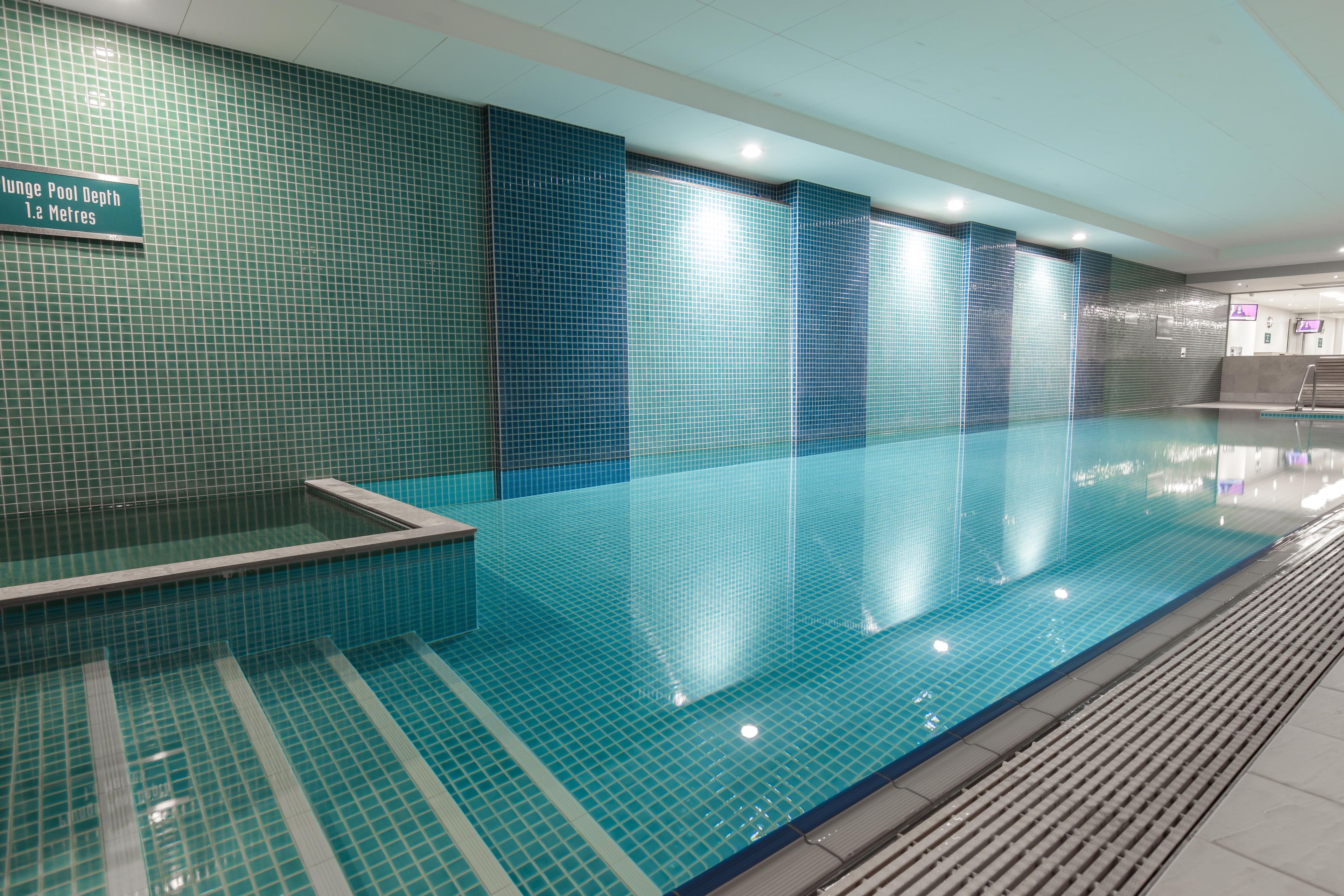 Una piscina interior con suelo y paredes de azulejos, un cartel que indica una profundidad de 1.2 metros y una zona de recepción con una pantalla de televisión.