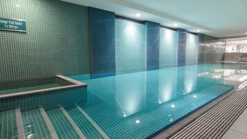 Una piscina interior con suelo y paredes de azulejos, un cartel que indica una profundidad de 1.2 metros y una zona de recepción con una pantalla de televisión.
