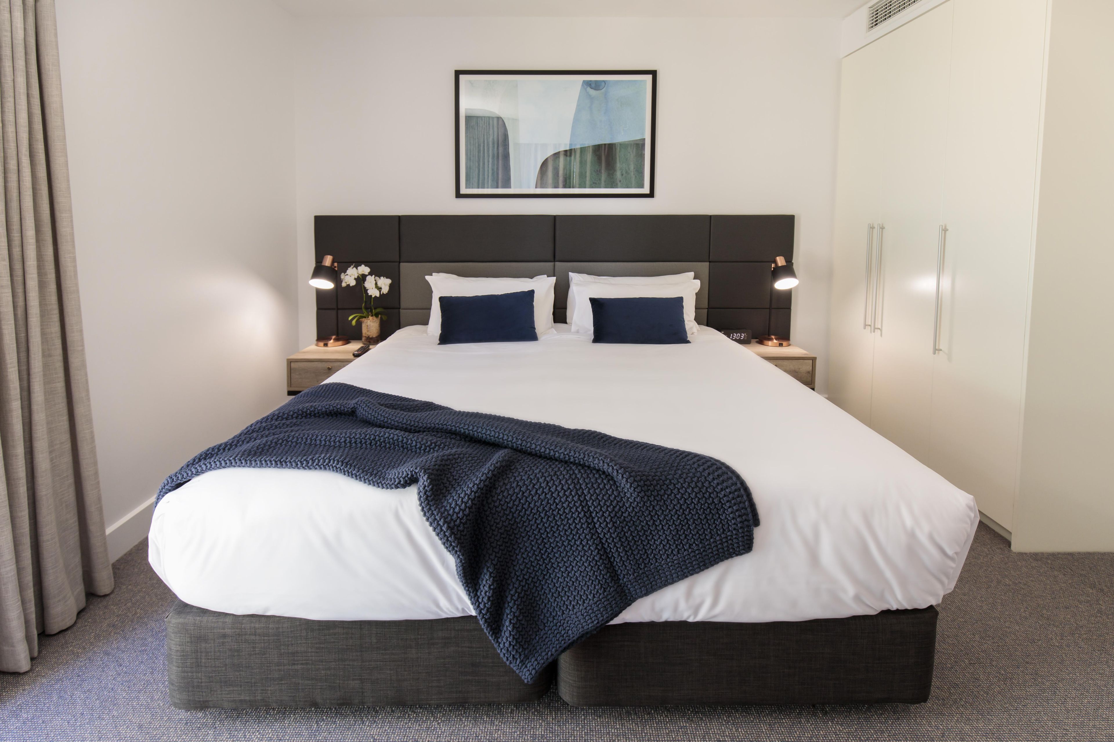 Habitación de hotel con una cama grande, dos mesitas de noche con lámparas, un cuadro en la pared y una ventana con cortinas.