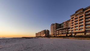 Un hôtel en bord de mer, avec plusieurs étages, des balcons et une plage de sable au premier plan.