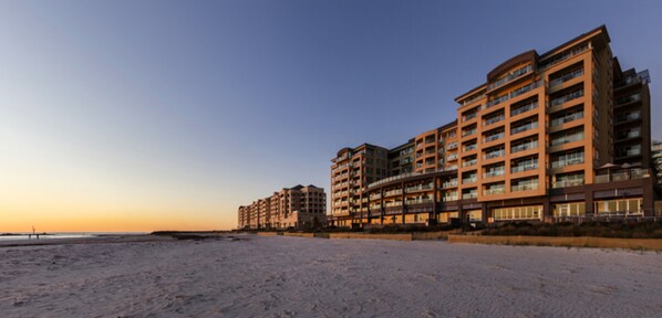 Un hôtel en bord de mer, avec plusieurs étages, des balcons et une plage de sable au premier plan.