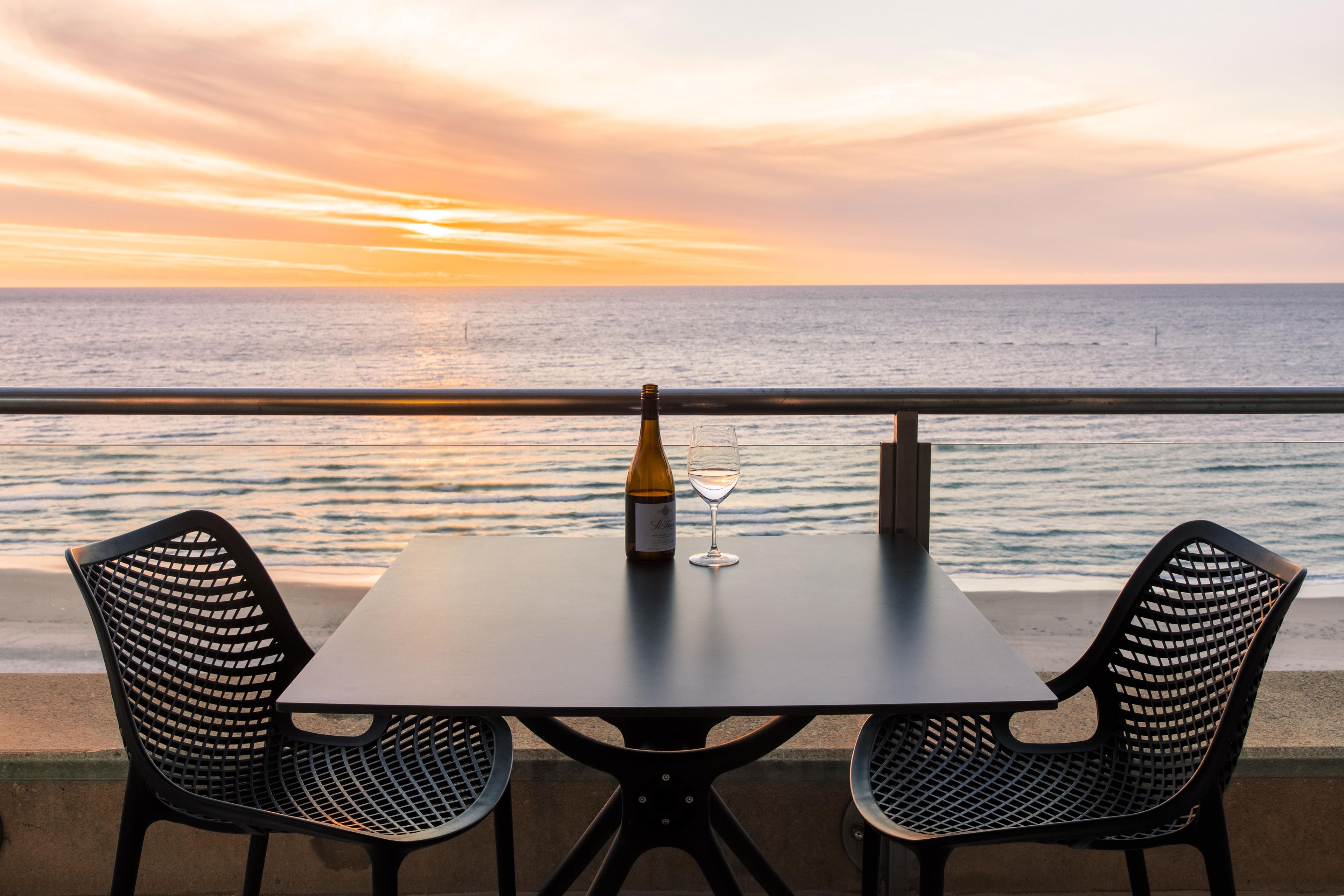 Une table avec deux chaises et une bouteille de vin, le tout placé devant un paysage marin illuminé par le coucher de soleil.