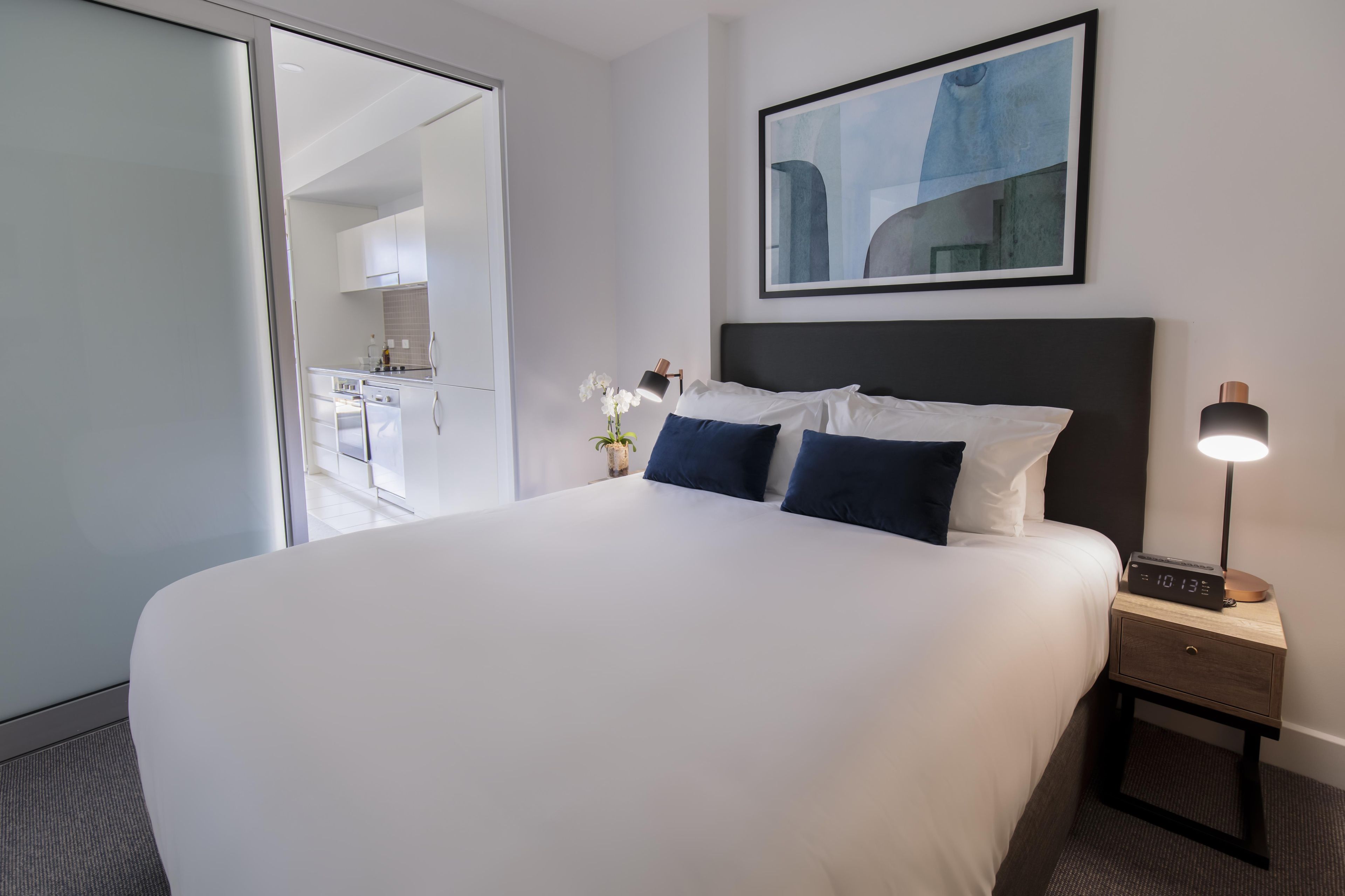 Una habitación de hotel con una cama grande, una mesita de noche con lámpara y vistas a la cocina.