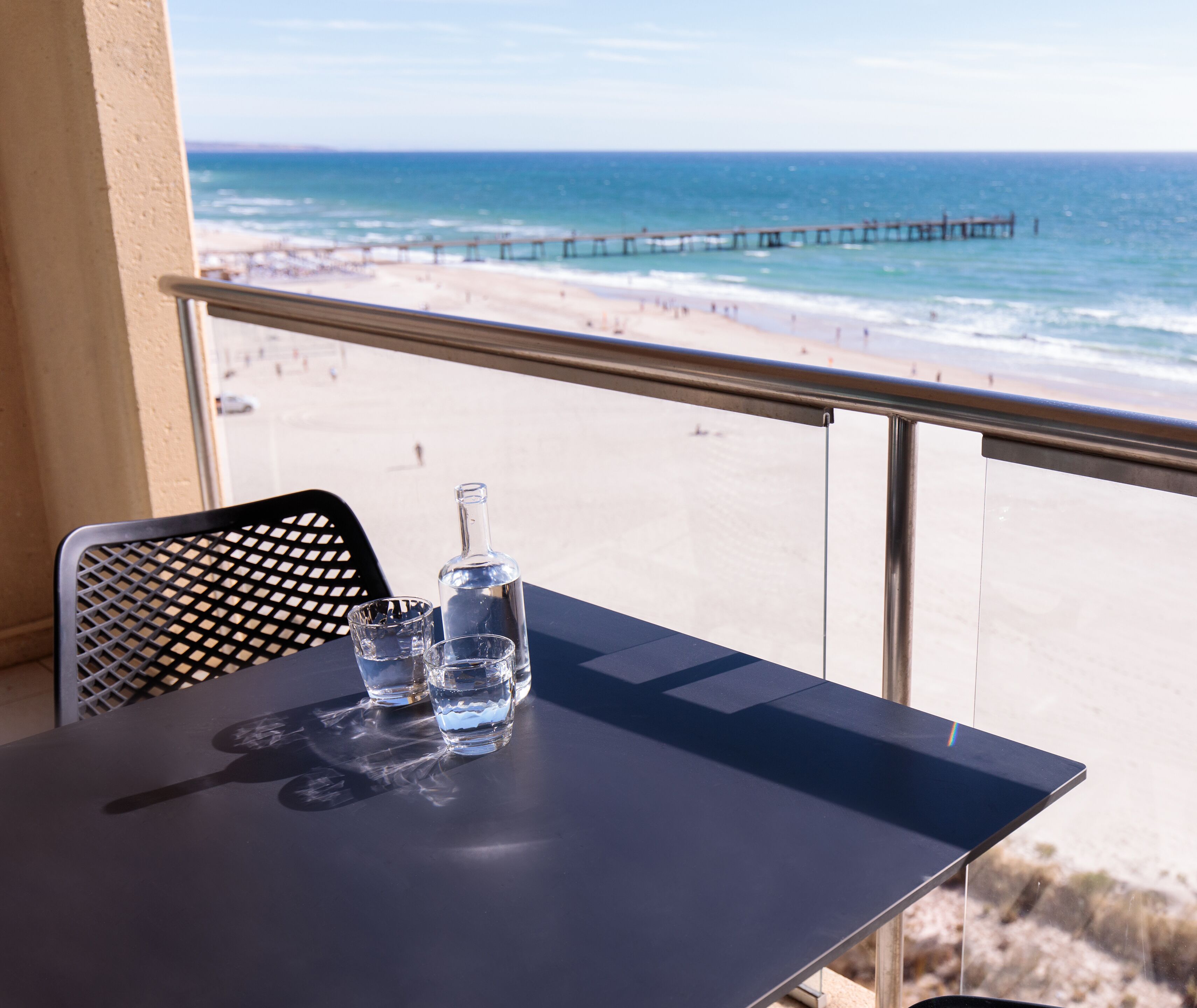 A balcony with a table, a bottle, and glasses. The view includes a beach and a pier.