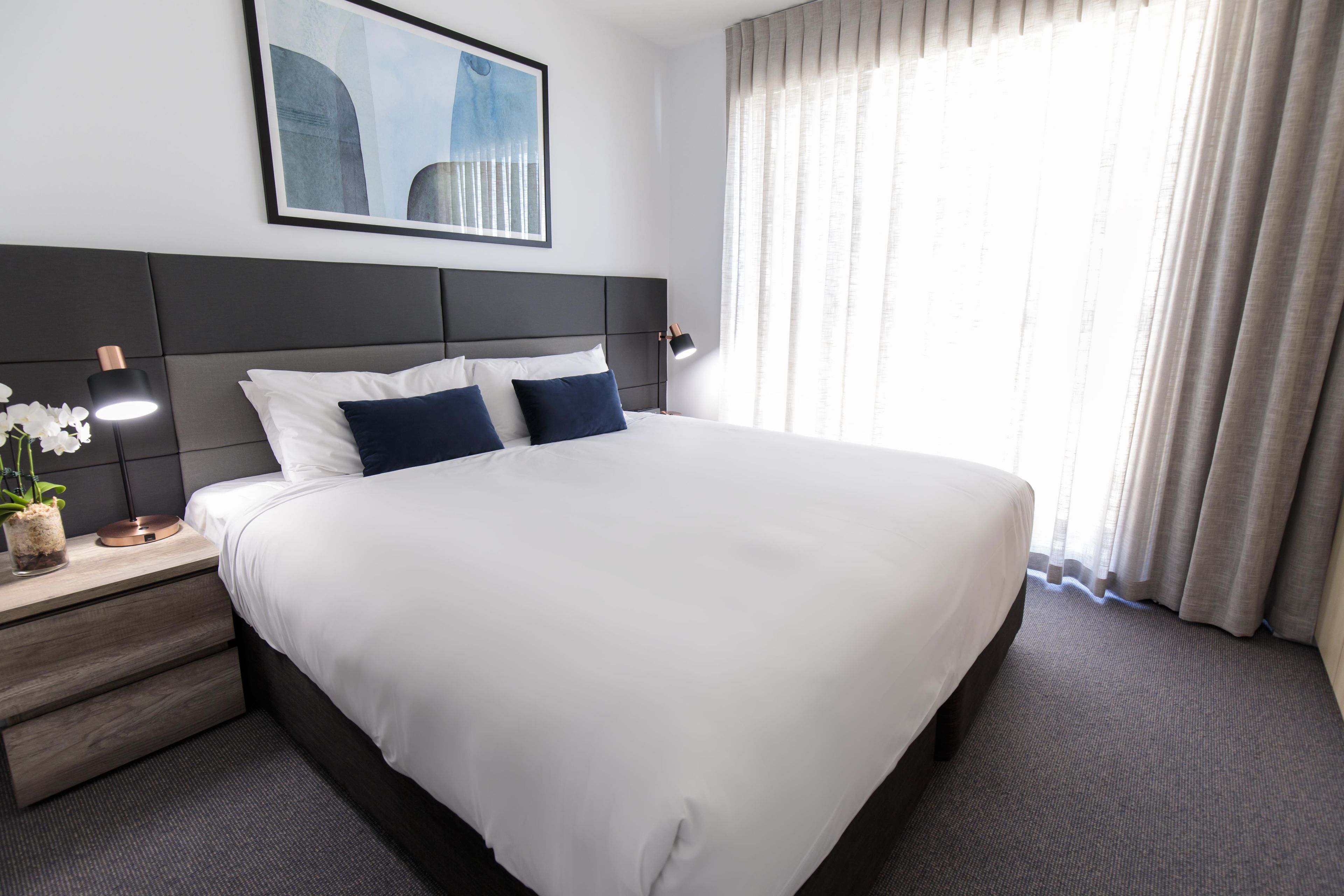 Ein Hotelzimmer mit einem großen Bett, einem Nachttisch mit Lampe, einem gerahmten Bild an der Wand und einem Fenster mit Vorhängen.