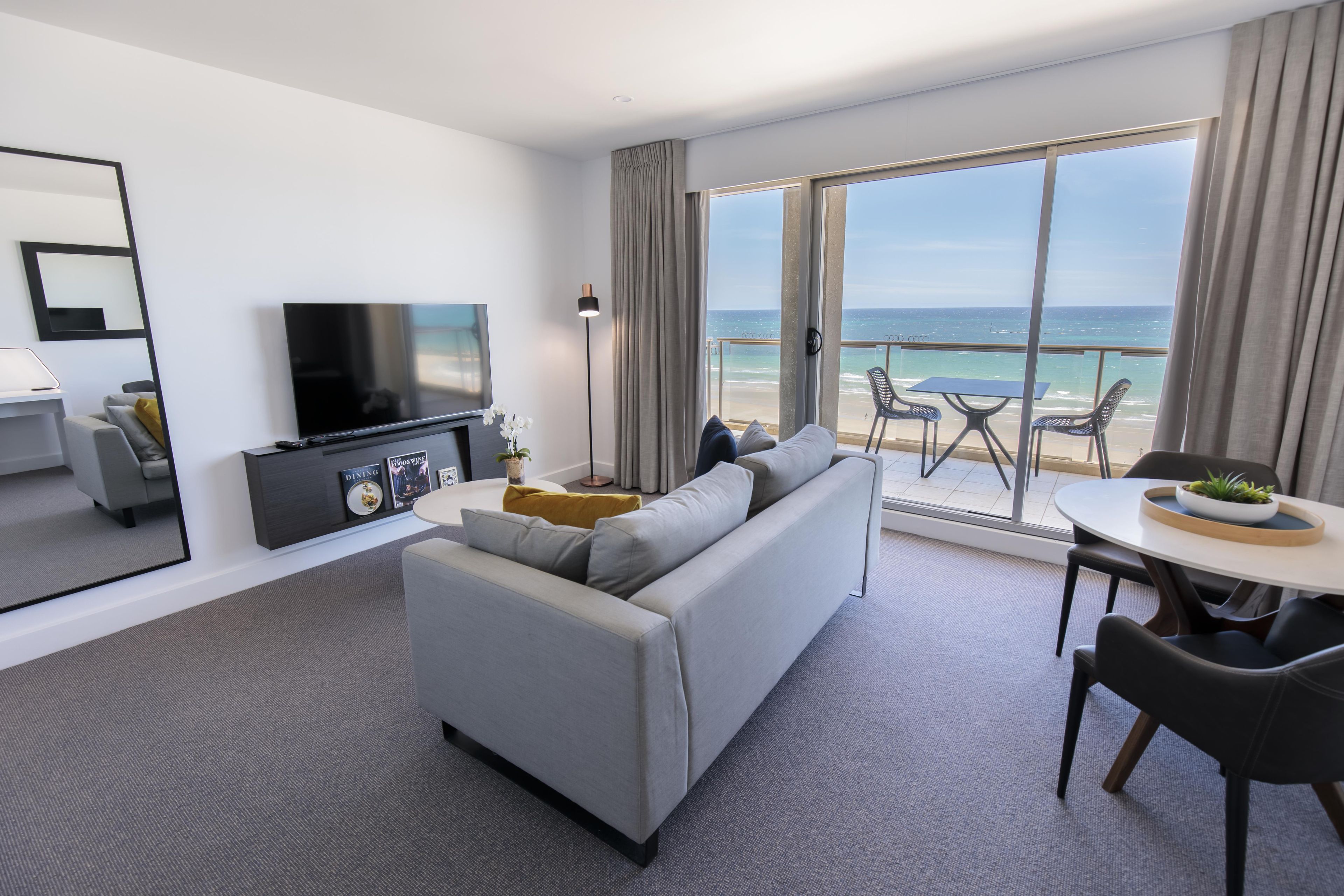 Quarto de hotel moderno com sofá cinzento, televisão de tela plana, área de refeições com mesa e cadeiras, e varanda com vista para o oceano.