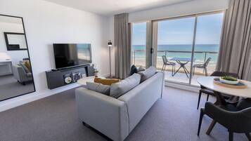 Quarto de hotel moderno com sofá cinzento, televisão de tela plana, área de refeições com mesa e cadeiras, e varanda com vista para o oceano.