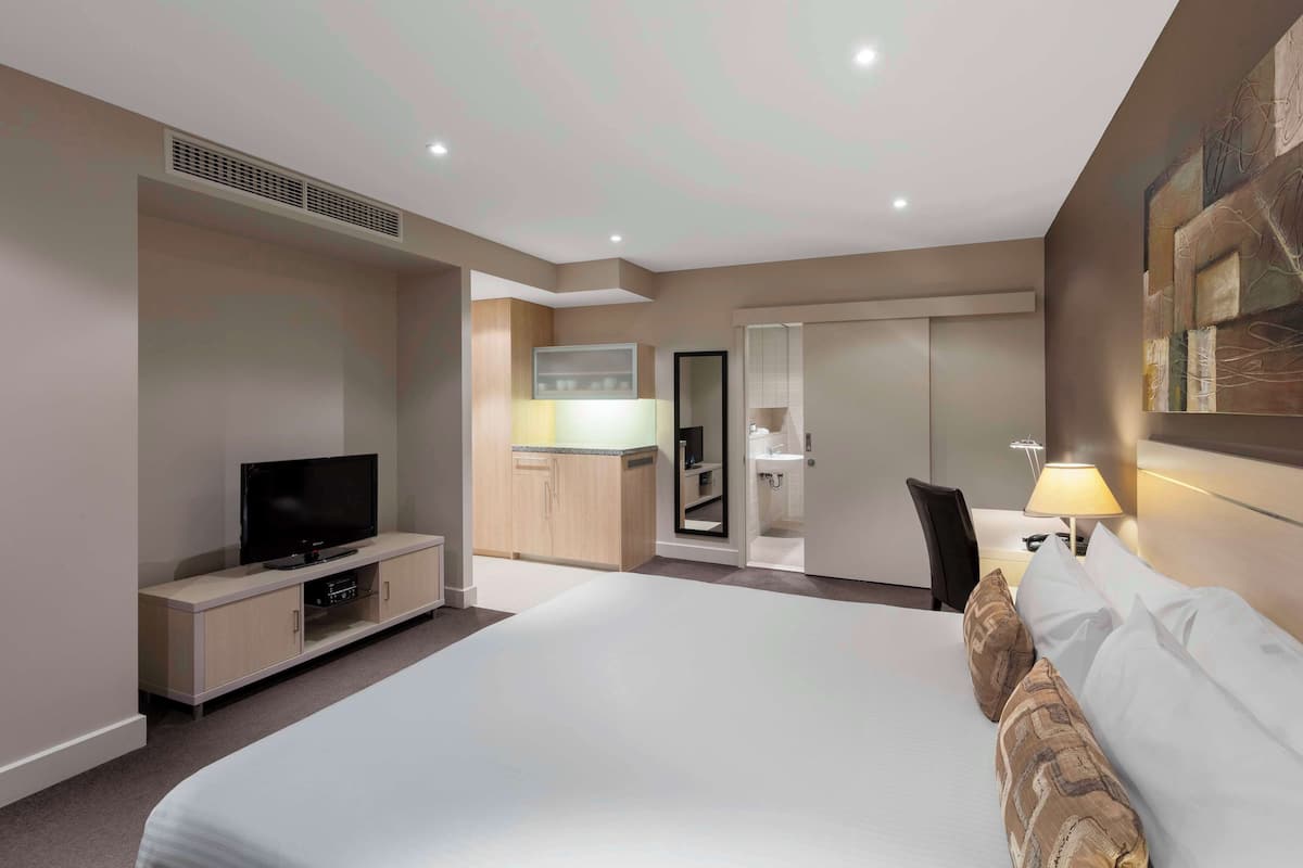 Hotelkamer met een groot bed, een televisie, een bureau met lamp en een kitchenette op de achtergrond.