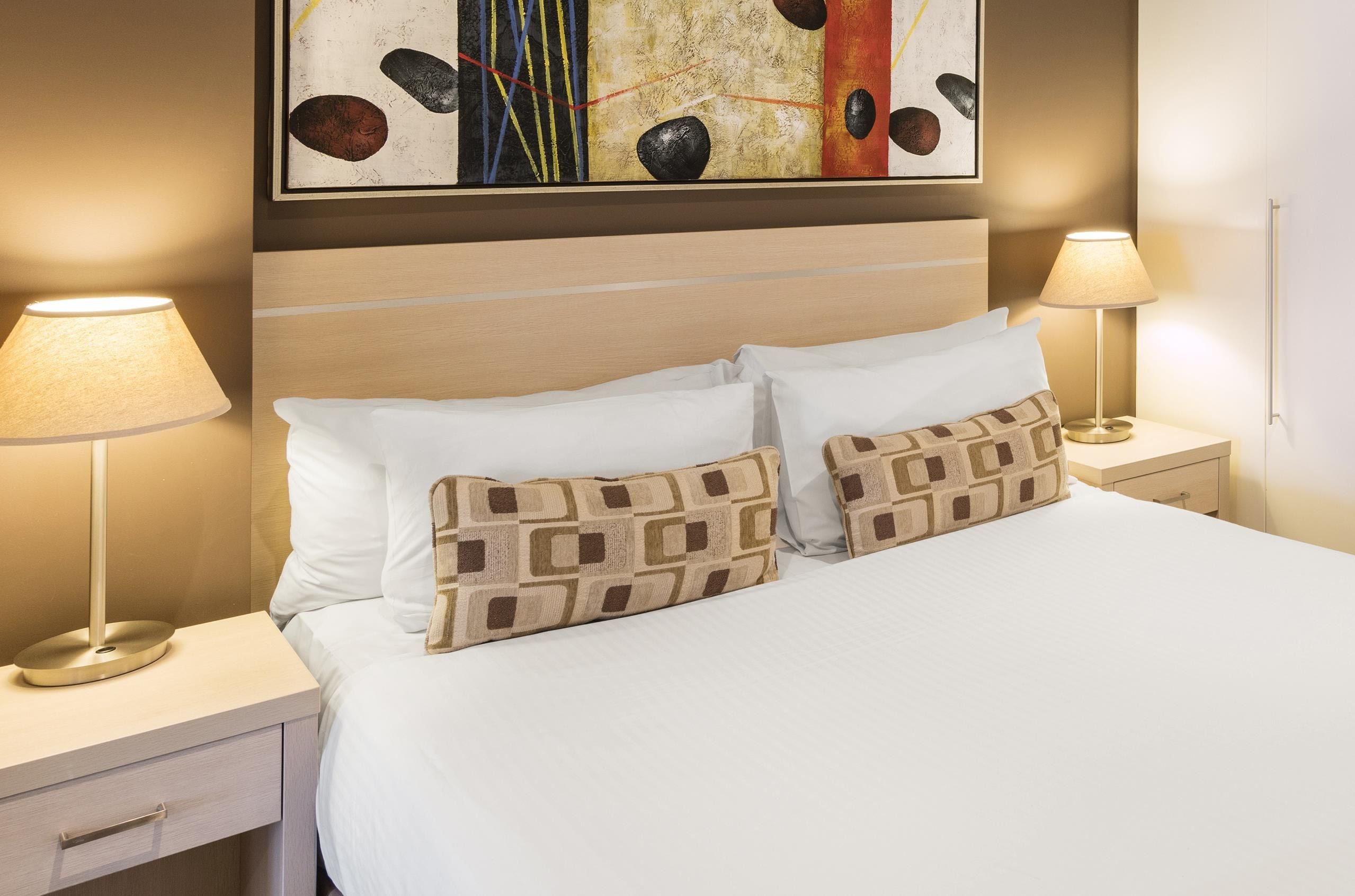A hotel room with a bed, two bedside lamps, a nightstand, a drawer, and a painting on the wall.