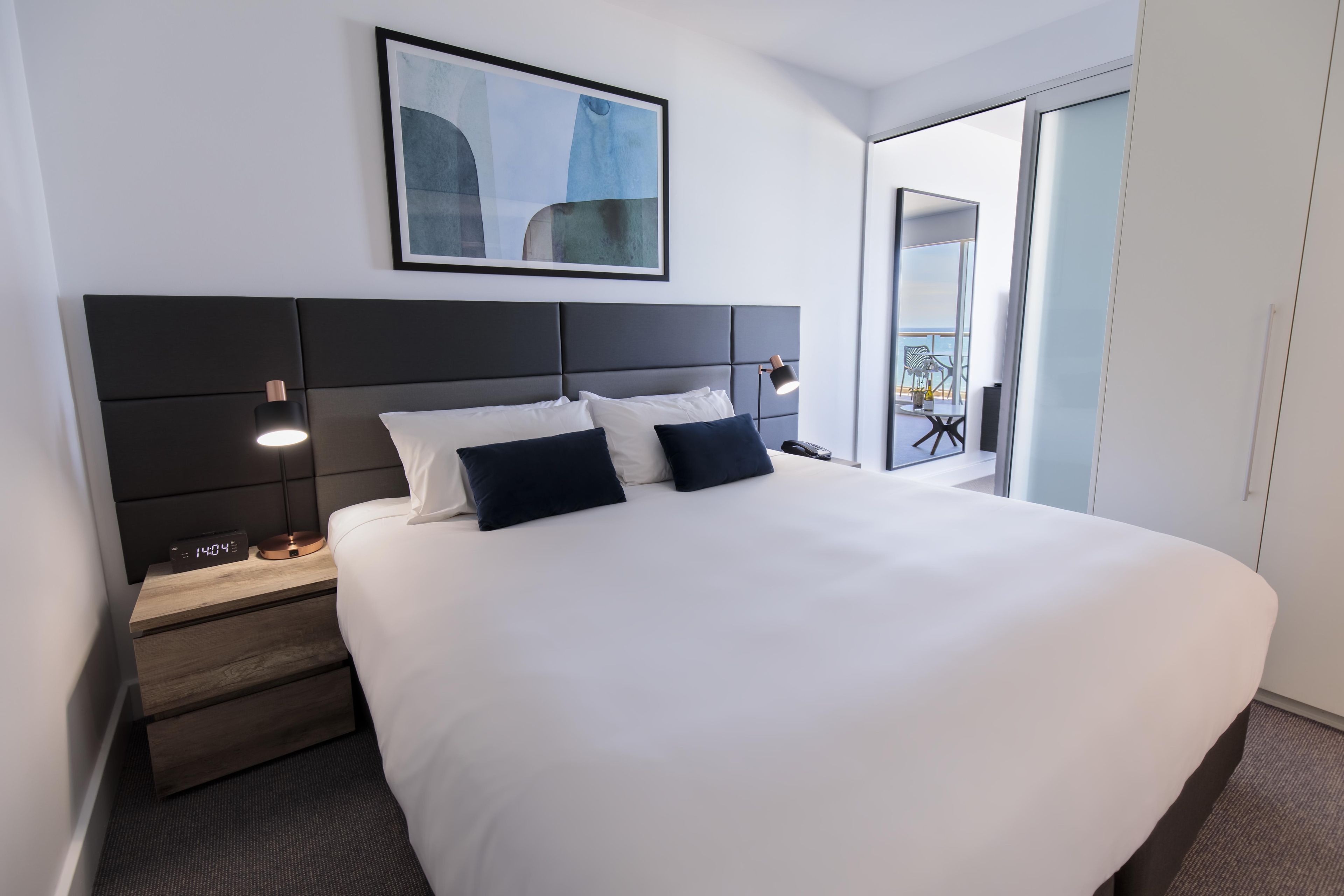 Habitación de hotel con una cama grande, una mesita de noche con lámpara, un cuadro enmarcado en la pared y vistas al exterior.