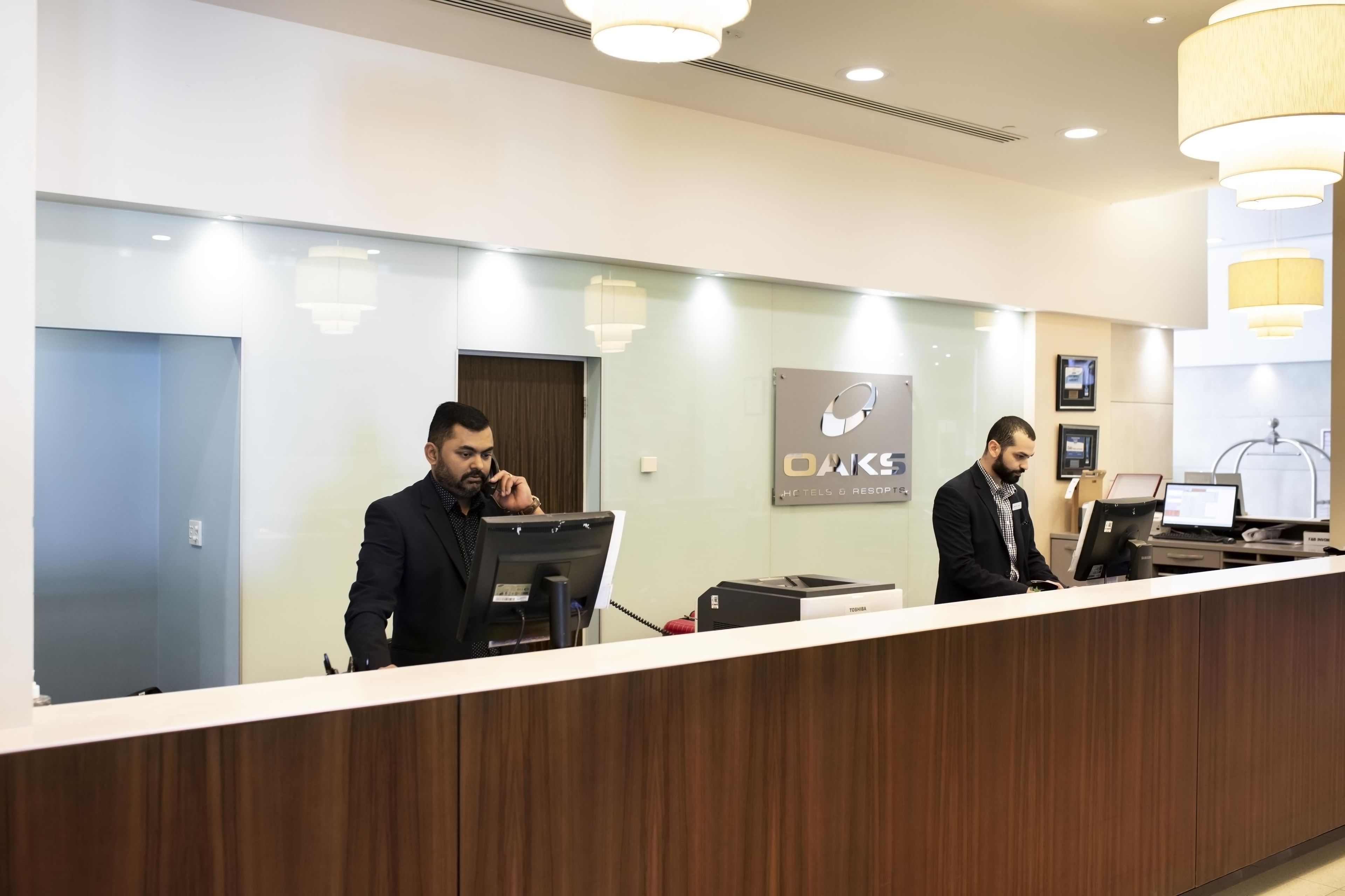 A reception desk with two staff members, a sign for OAKS Hotels & Resorts, and a computer.