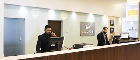 A reception desk with two staff members, a sign for OAKS Hotels & Resorts, and a computer.