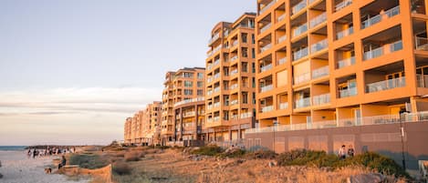 A beachfront hotel with multiple balconies, a sandy beach in the foreground, and a clear sky.