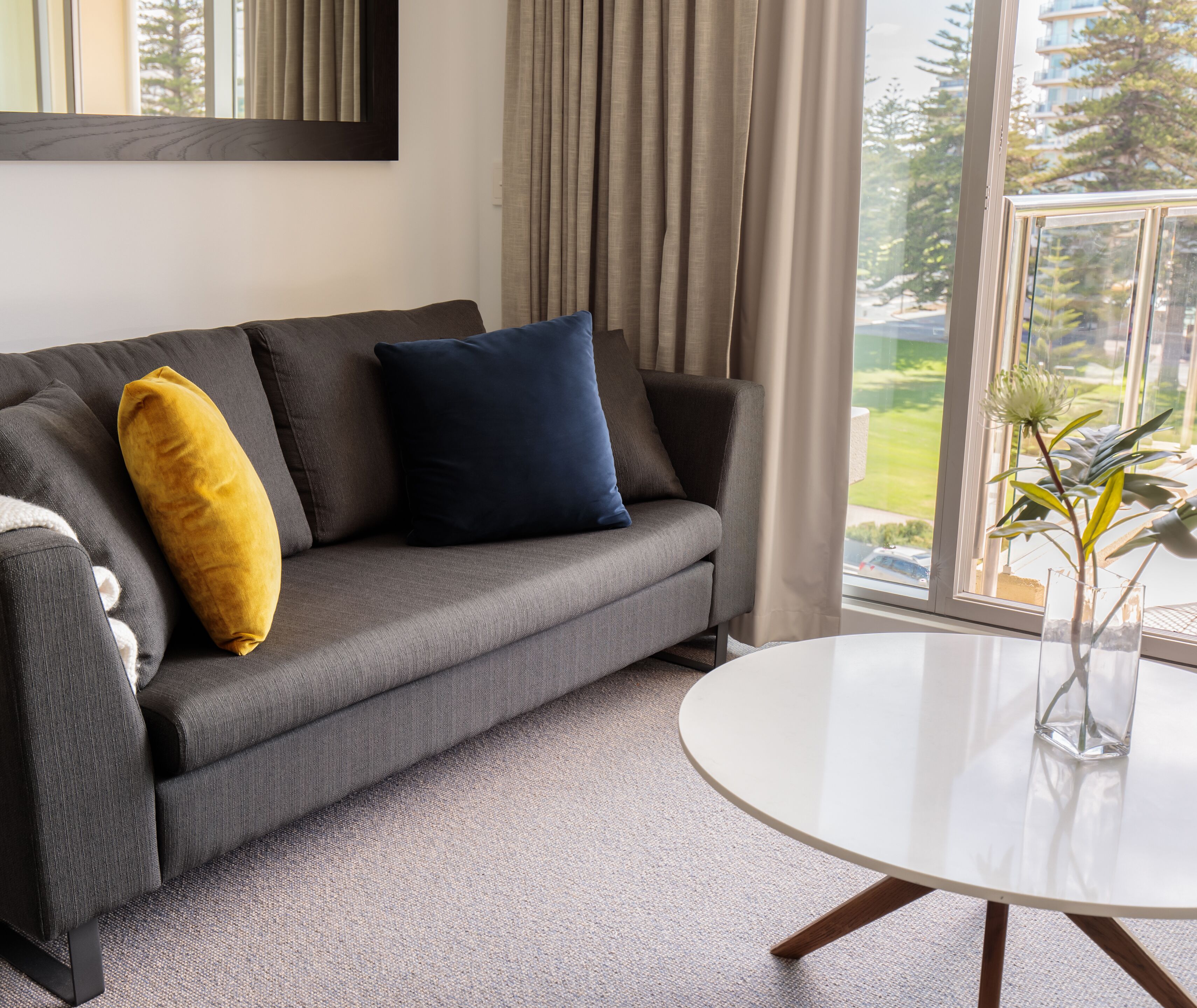 Una sala de estar moderna con un sofá gris, una almohada amarilla y otra azul, una mesa redonda blanca y vista a un paisaje verde a través de amplias ventanas.
