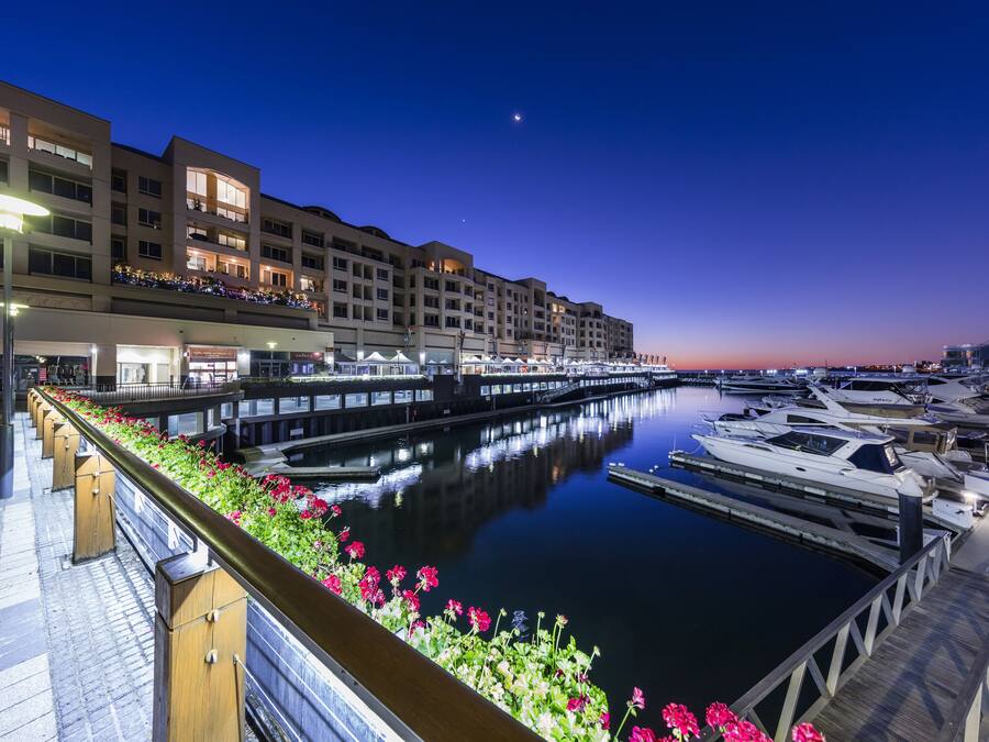 A marina with boats docked, a waterfront promenade, and a building with balconies overlooking the water.
